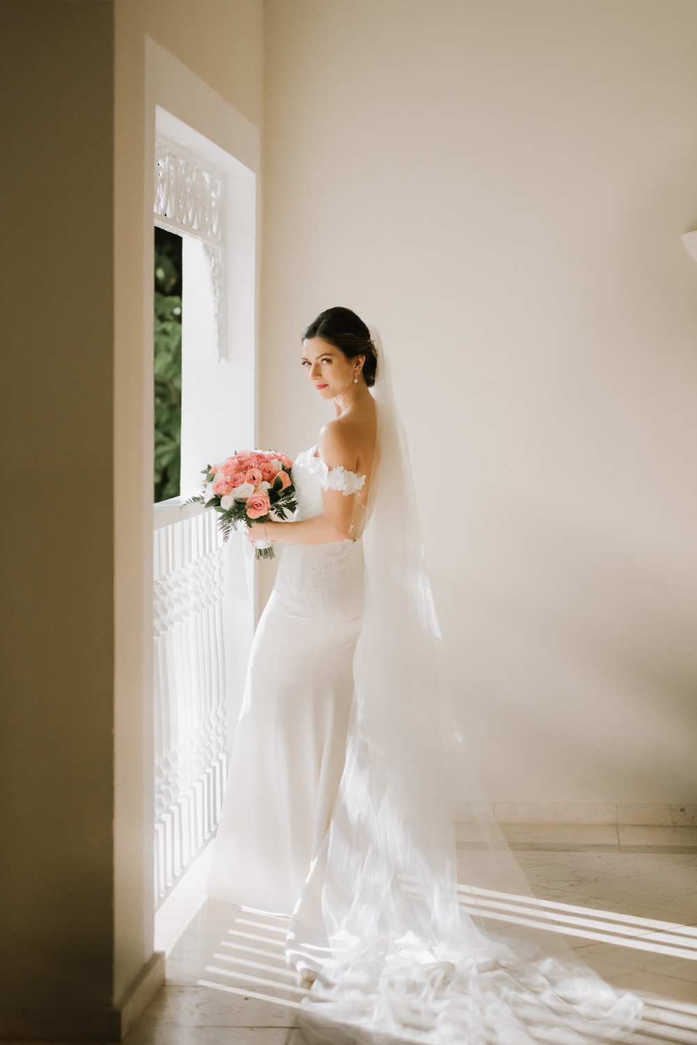 The bride in a wedding dress, veil, with a bouquet of white flowers is standing by the window.