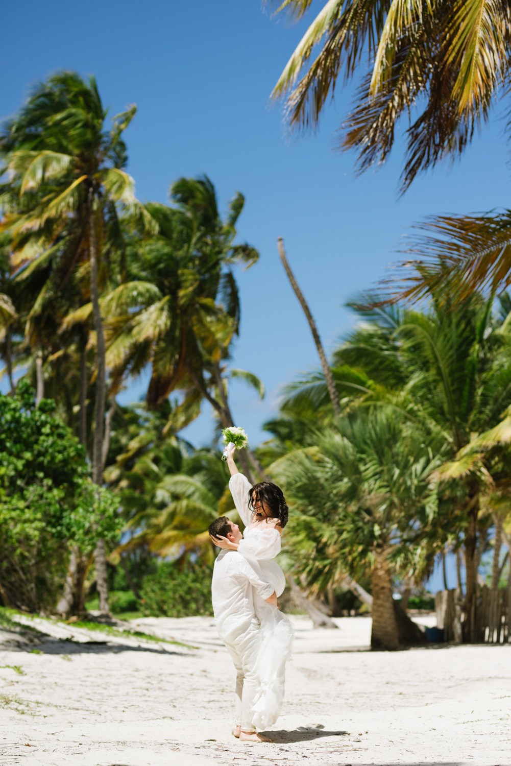 The groom lifts the bride up on the beach with palm trees.