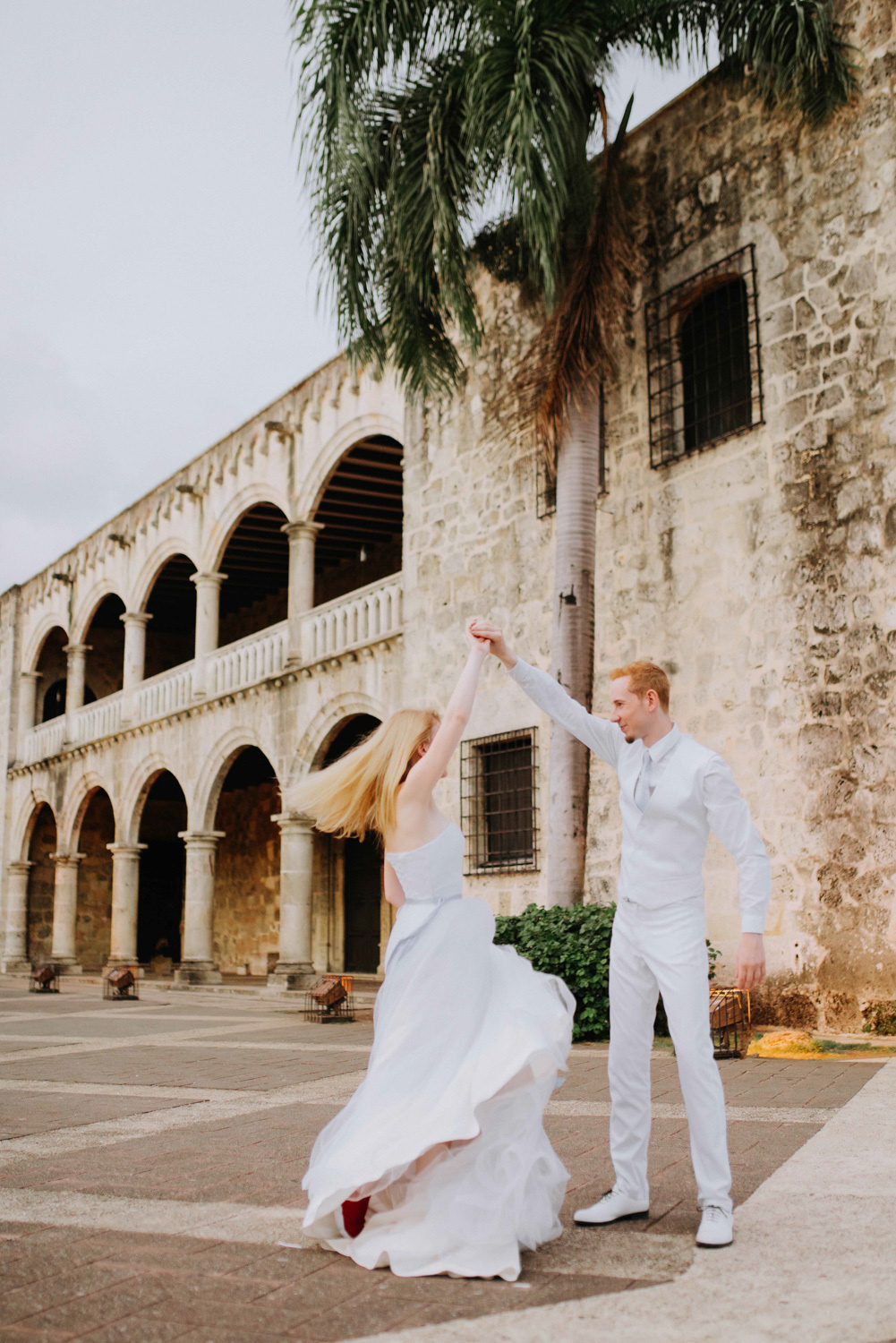 Невеста кружится, держа за руку жениха, на фоне старинного здания и пальмы.