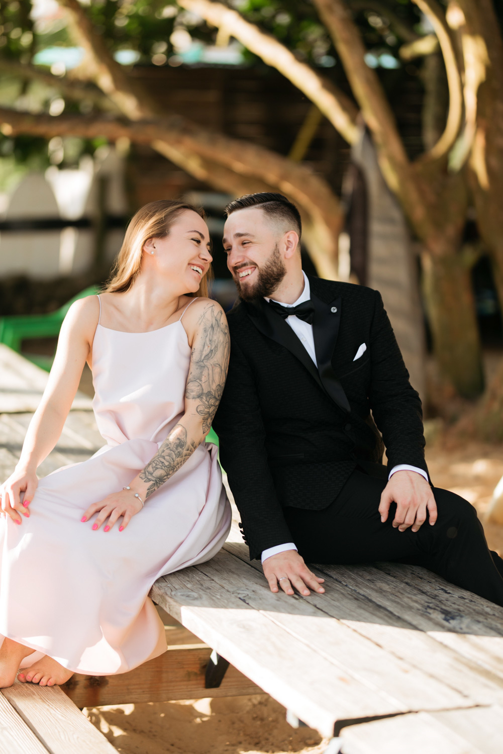 The bride and groom are sitting on a wooden bench under a tree.