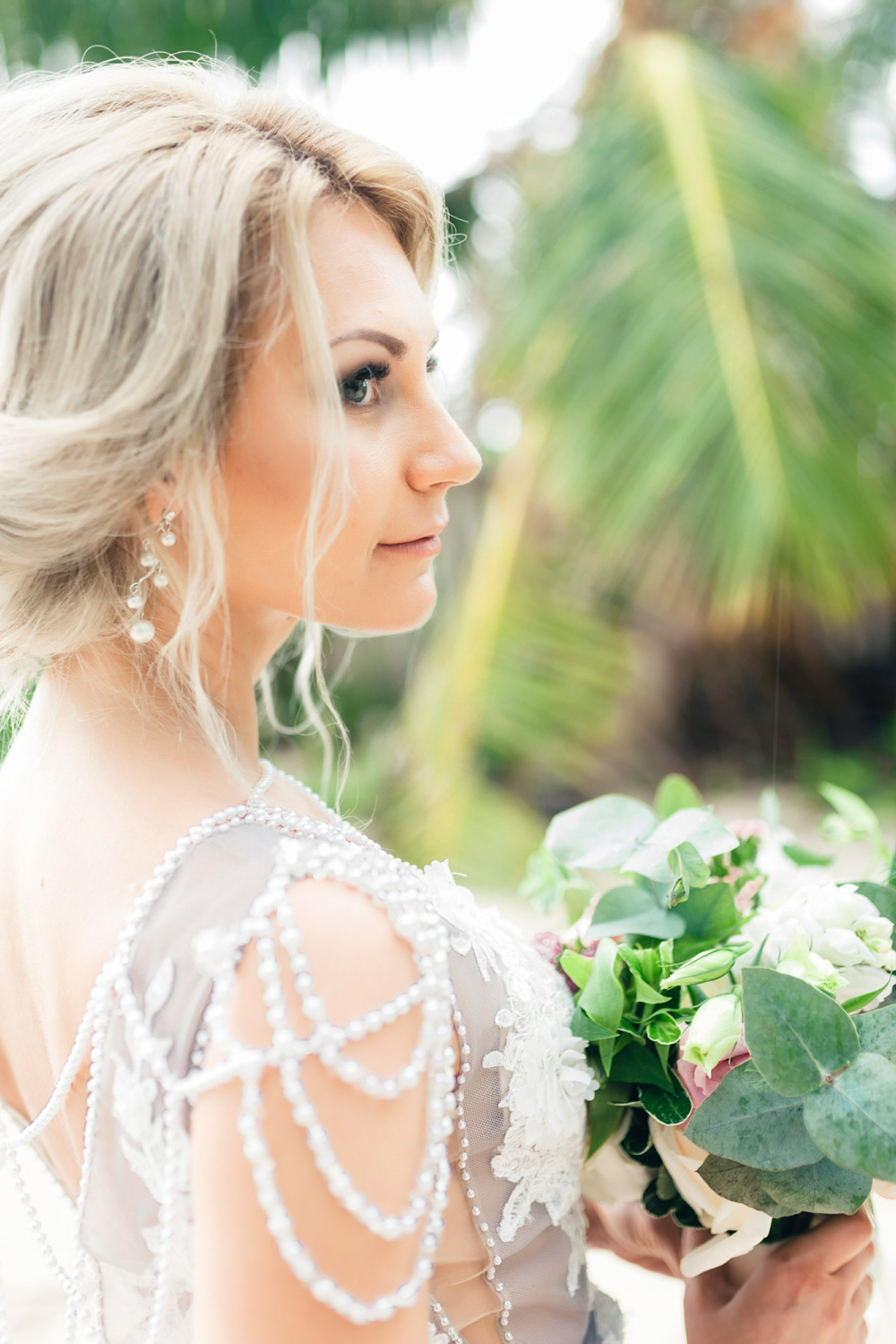 The bride's face with a beautiful wedding hairstyle and a bouquet of flowers in her hands on the background of a palm tree.