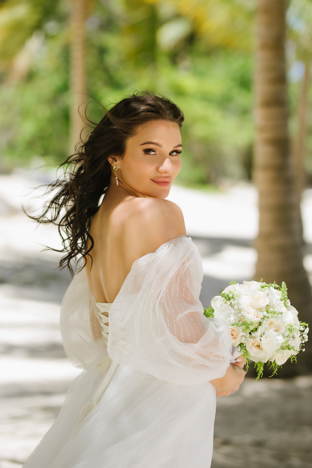 A bride in an open-back wedding dress, turning around, looks back at the background of palm trees.