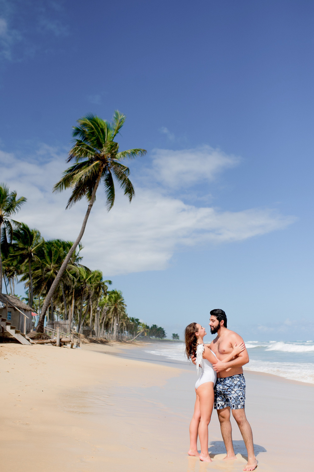 The bride in a white solid swimsuit and the groom in gray and white flowered shorts look at each other, holding hands, against the background of the sea beach with palm trees.