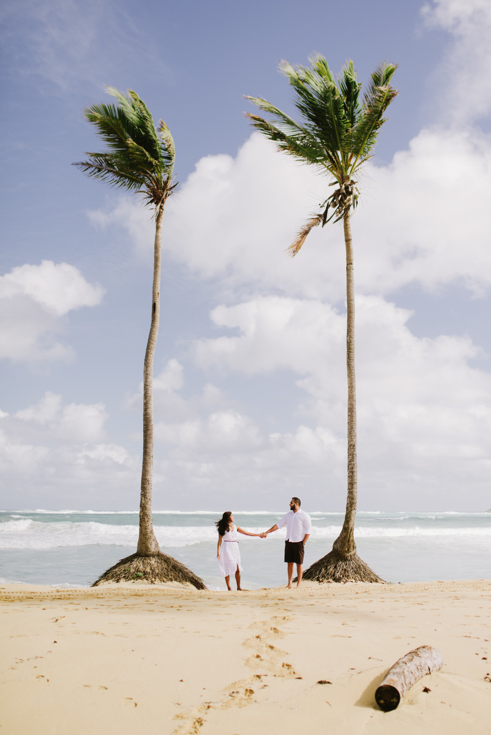 Жених с невестой стоят между двух пальм на фоне моря, взявшись за руку, оставив позади себя следы на песке.