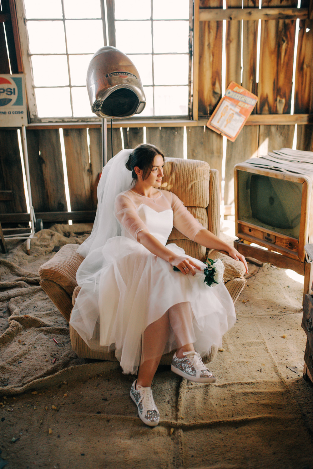 Невеста сидит в старом деревянном помещении в старинном кресле рядом со старым телевизором. У неё за спиной старинная сушилка для волос.