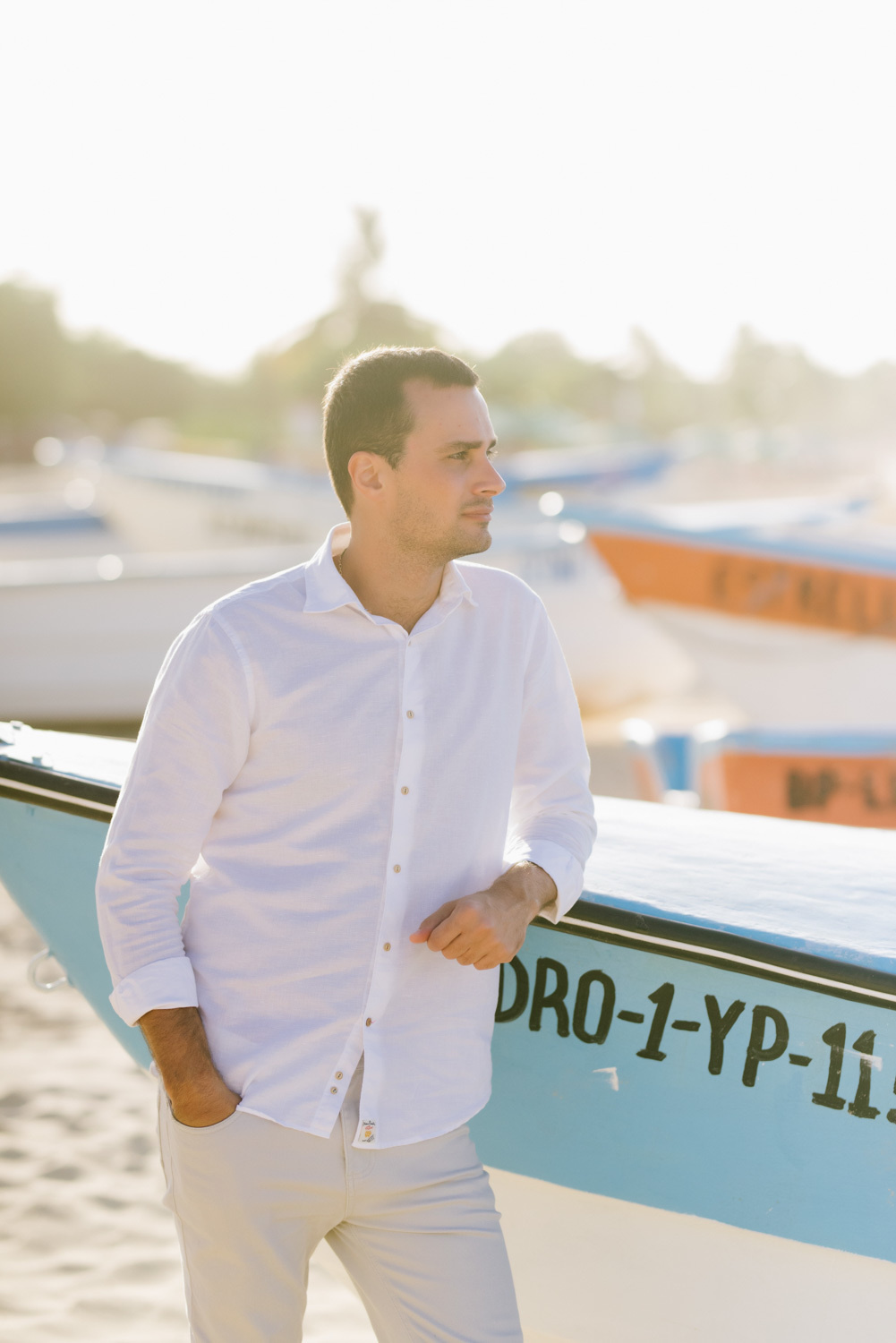 a man (groom) in a white shirt and light trousers poses by a wooden boat on the beach