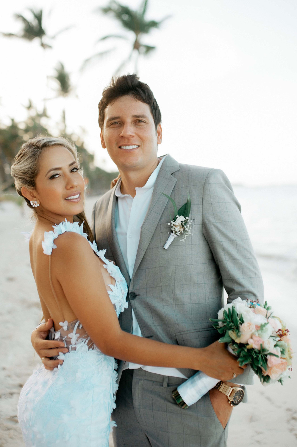 Bride and groom hug each other on the background of the sea beach. The bride is holding a bouquet of flowers.