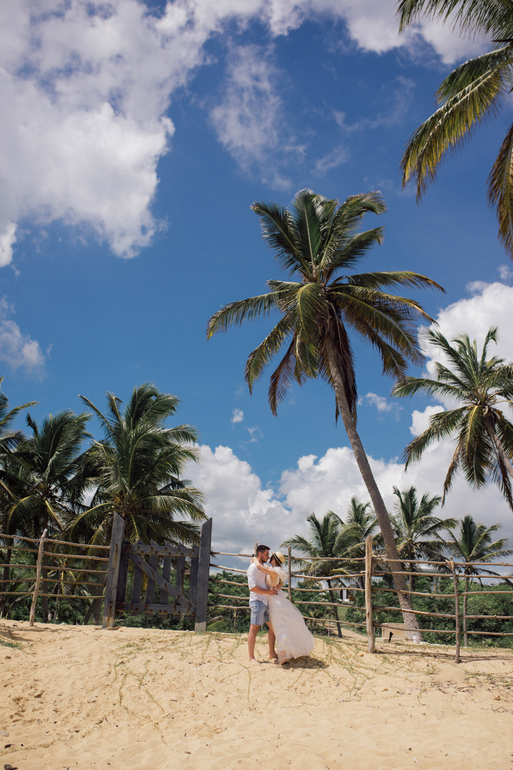 On the sand, the bride and groom stand with their arms around each other against the background of a wooden fence and palm trees.