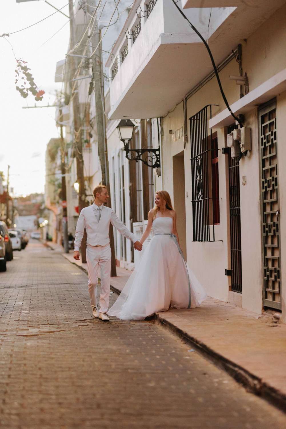 Жених с невестой, взявшись за руки, идут по улице города.