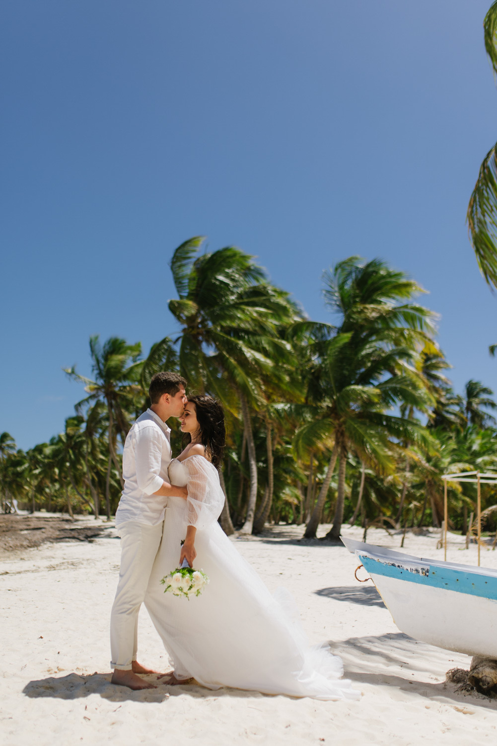 The groom kisses the bride on the forehead at the boat on the beach with palm trees.