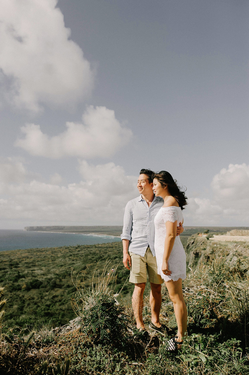 On the green grass, a dark-haired girl with long fluffed hair in a short white dress and a dark-haired guy with short hair in a light shirt and light shorts are standing on the green grass against the blue sky and looking at the sea.