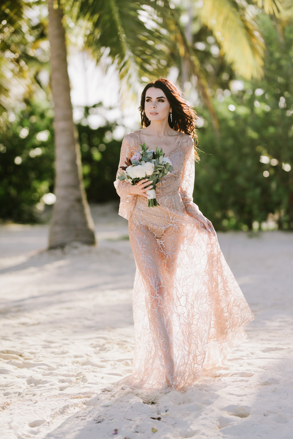 Невеста в розовом платье с букетом цветов на фоне пальм.