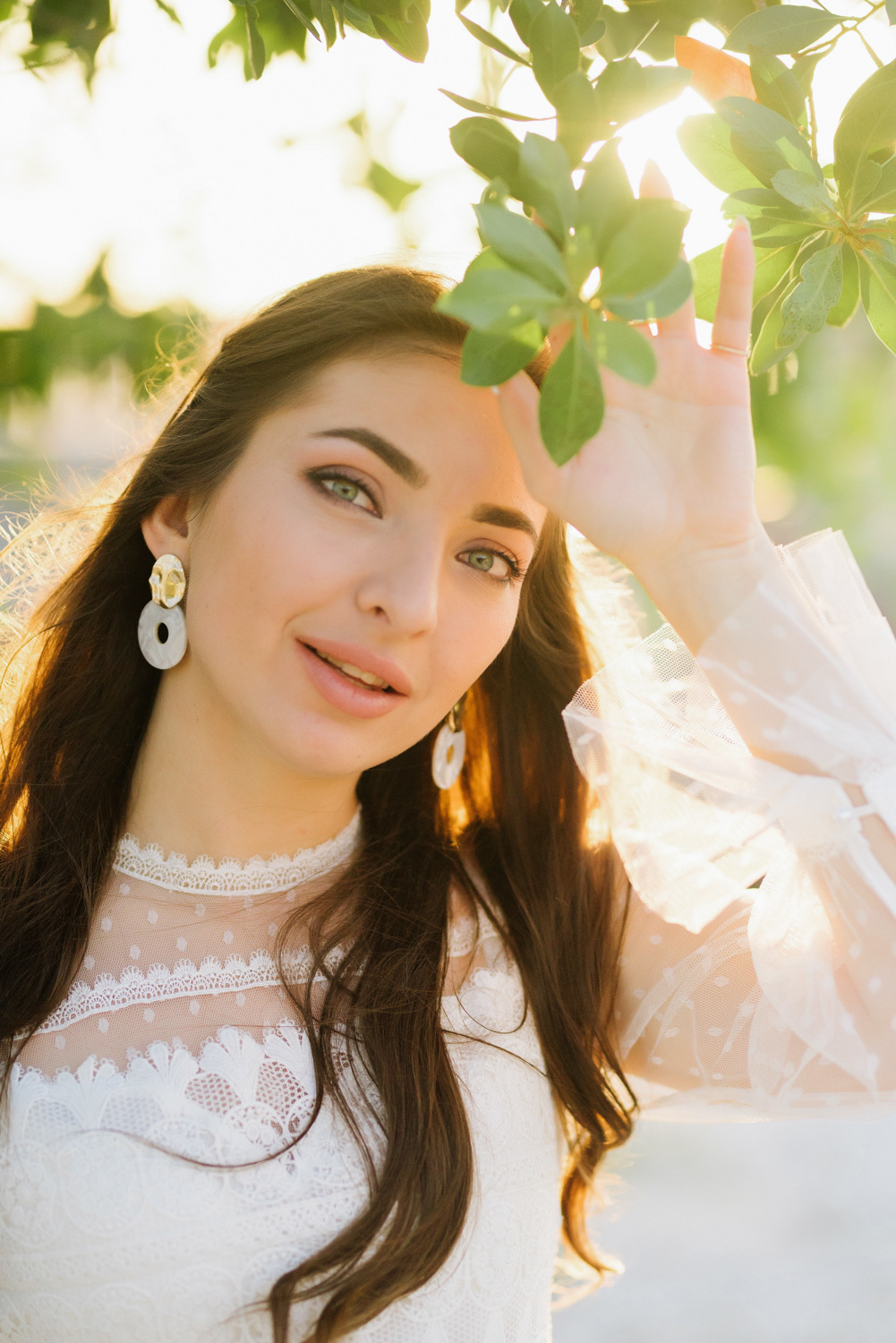 The bride's face with loose brown hair, white earrings on the background of a tree branch.