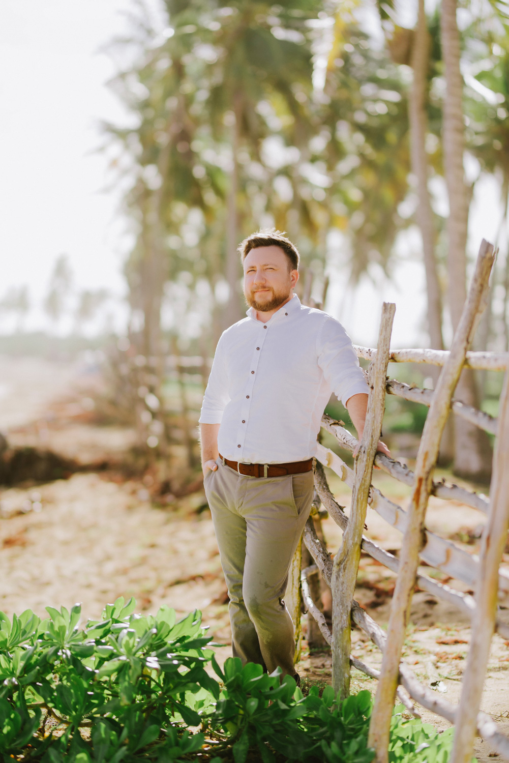 The groom stands at a wooden fence against a background of palm trees.