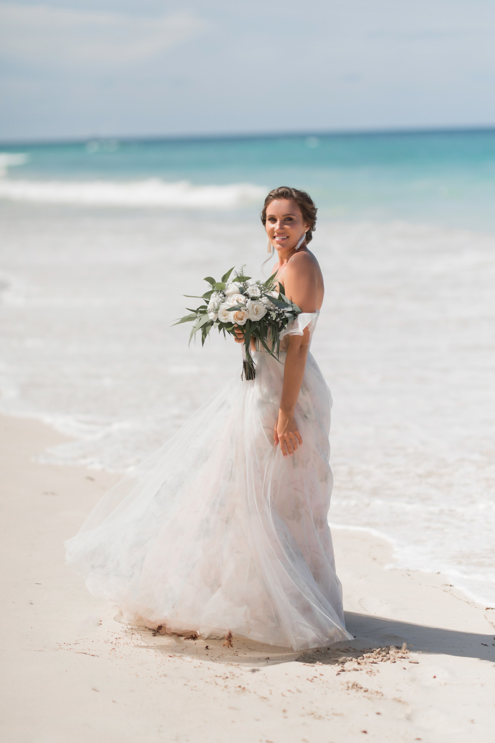 Невеста в развевающемся лёгком платье с букетом цветов на фоне морских волн.