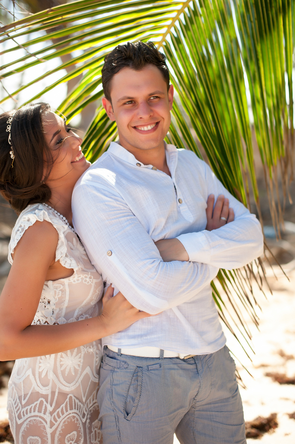 The groom in a white shirt and gray shorts and the bride in a white openwork dress on the background of palm leaves.