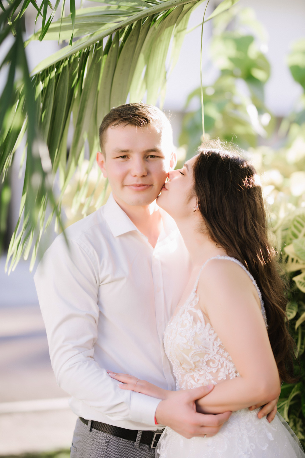 A girl in an openwork white dress kisses the groom in a white shirt under the leaves of a palm tree.