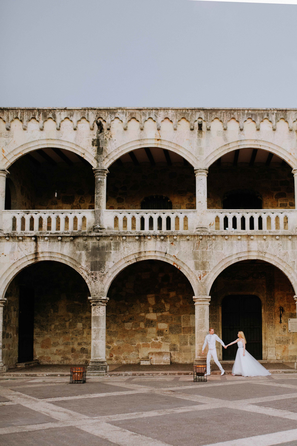 Жених ведёт невесту за руку вдоль высокого серого здания с двухъярусными арками.