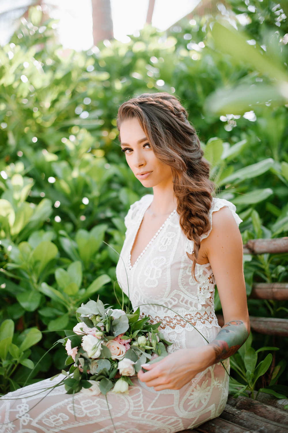 The bride is sitting on a bench with a bouquet of flowers on the background of a green shrub.