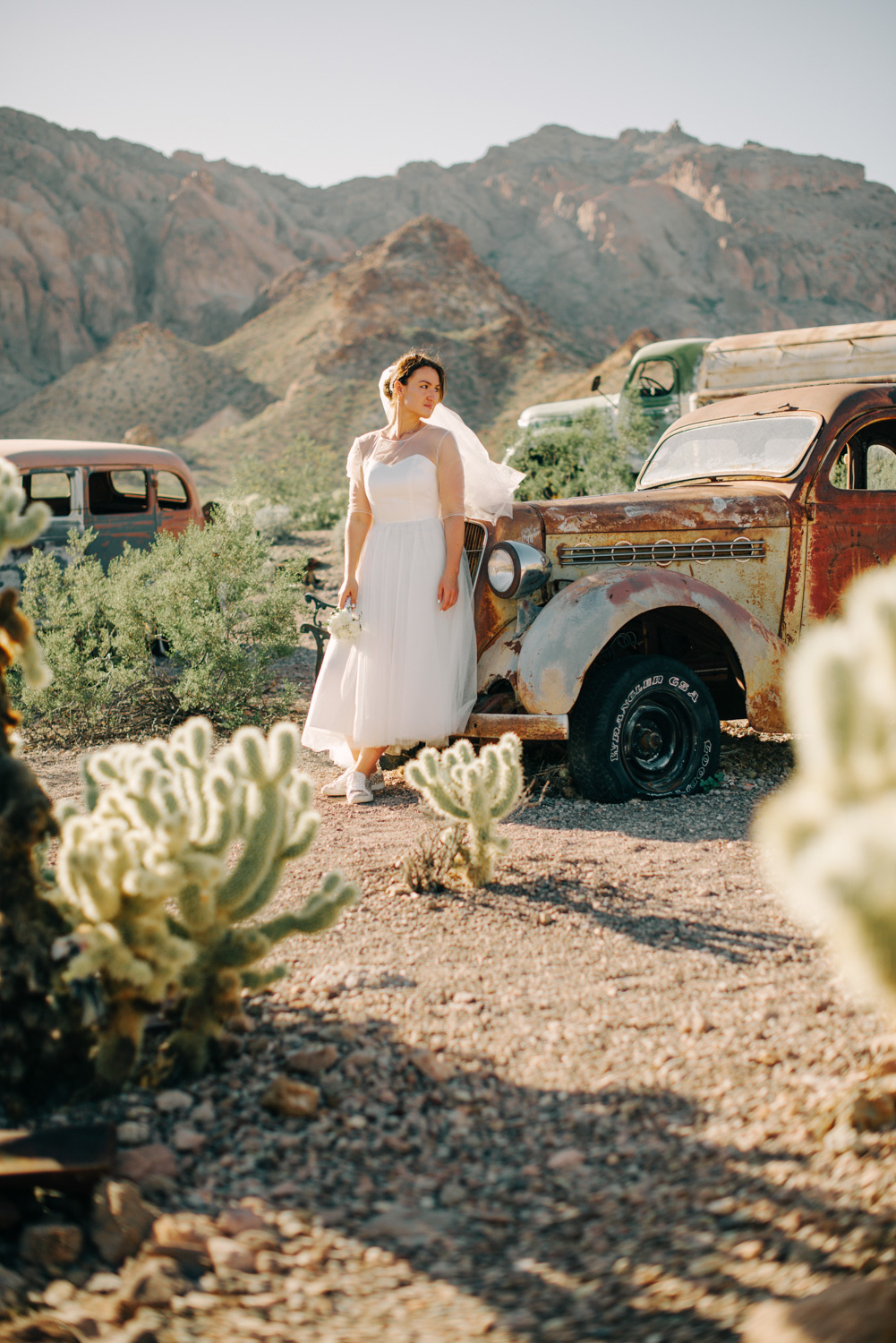 Невеста стоит среди заброшенных машин, кактусов на фоне гор.