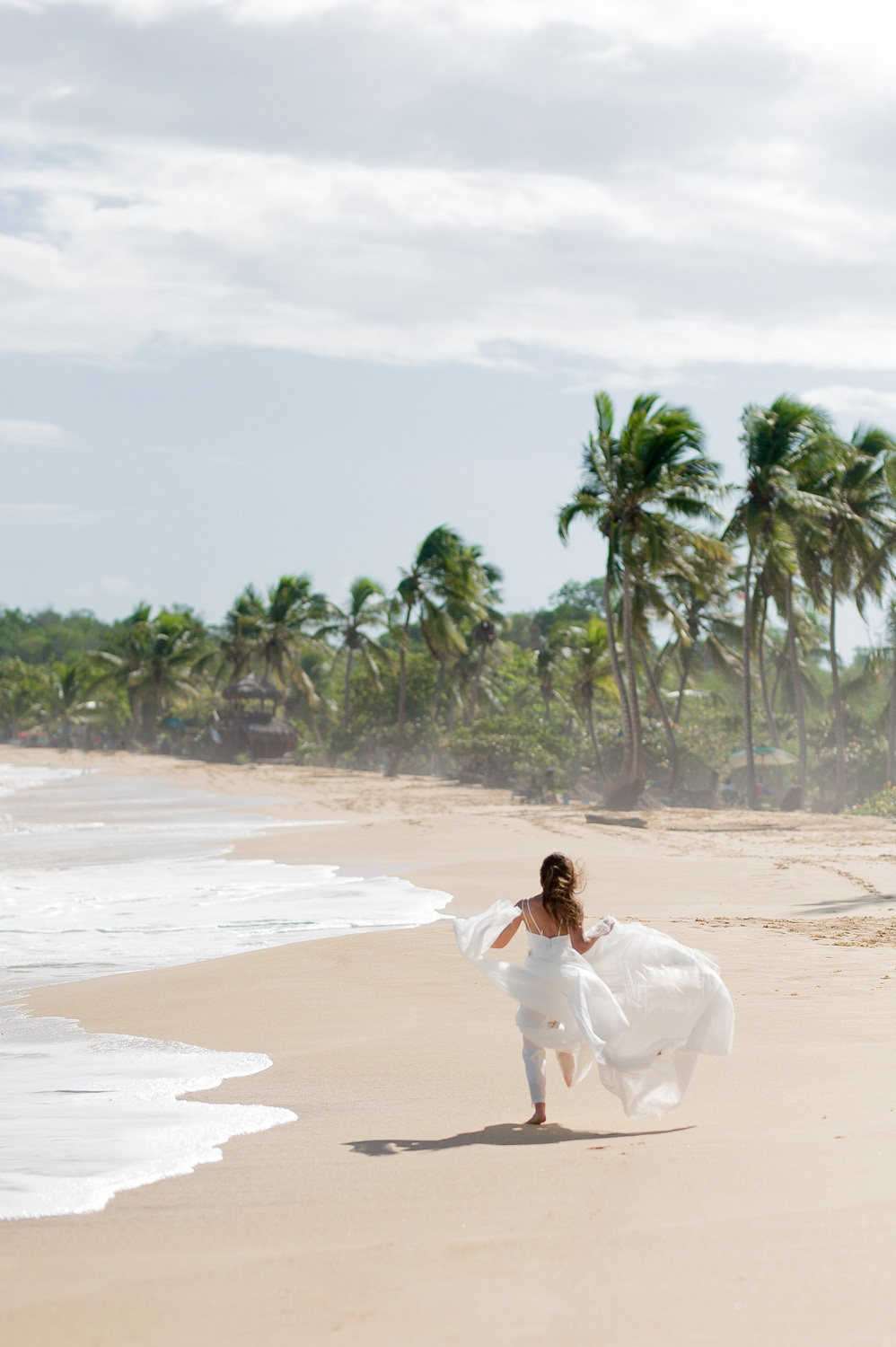 The bride runs along the sandy beach against the background of sea foam and palm trees along the coast.