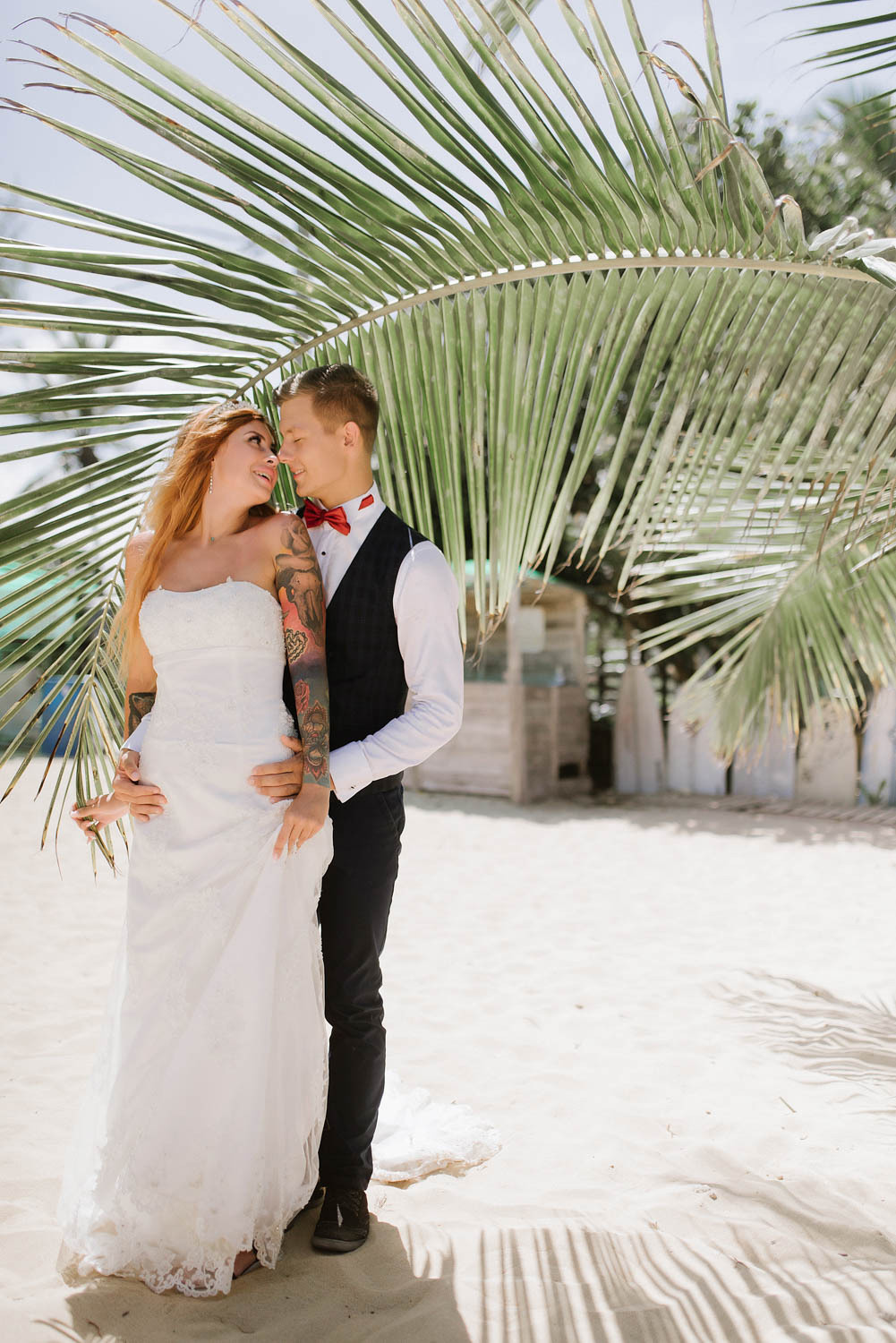On a sandy beach, the groom embraces the bride against the background of a palm leaf.