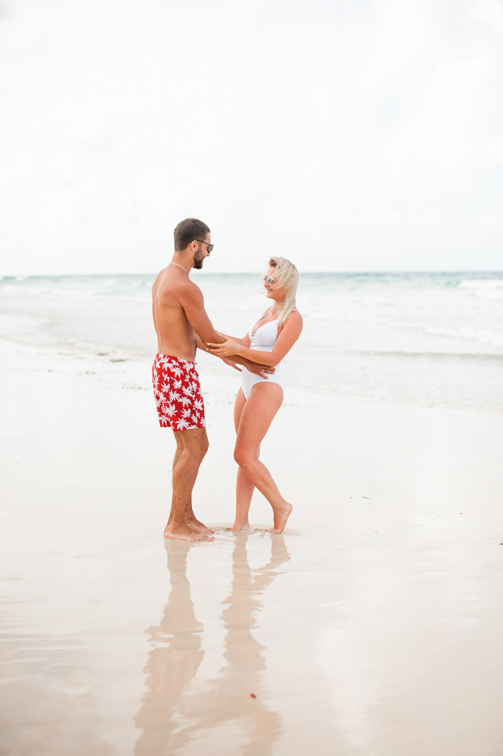The groom in red and white underpants and the bride in a solid white swimsuit hold hands against the background of the sea.