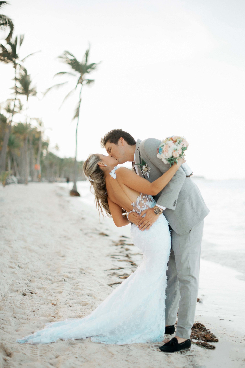 The groom, bent low, kisses the bride on the background of the beach with palm trees and the sea.