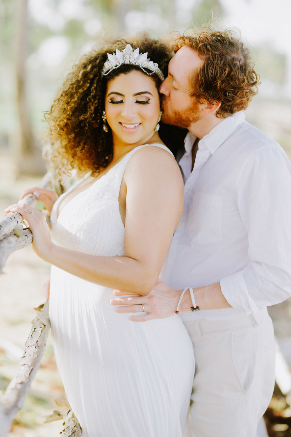 Невесту в свадебном платье обнимает жених с вьющимися каштановыми волосами и бородой в белой рубашке и брюках на расплывчатом фоне пальм.