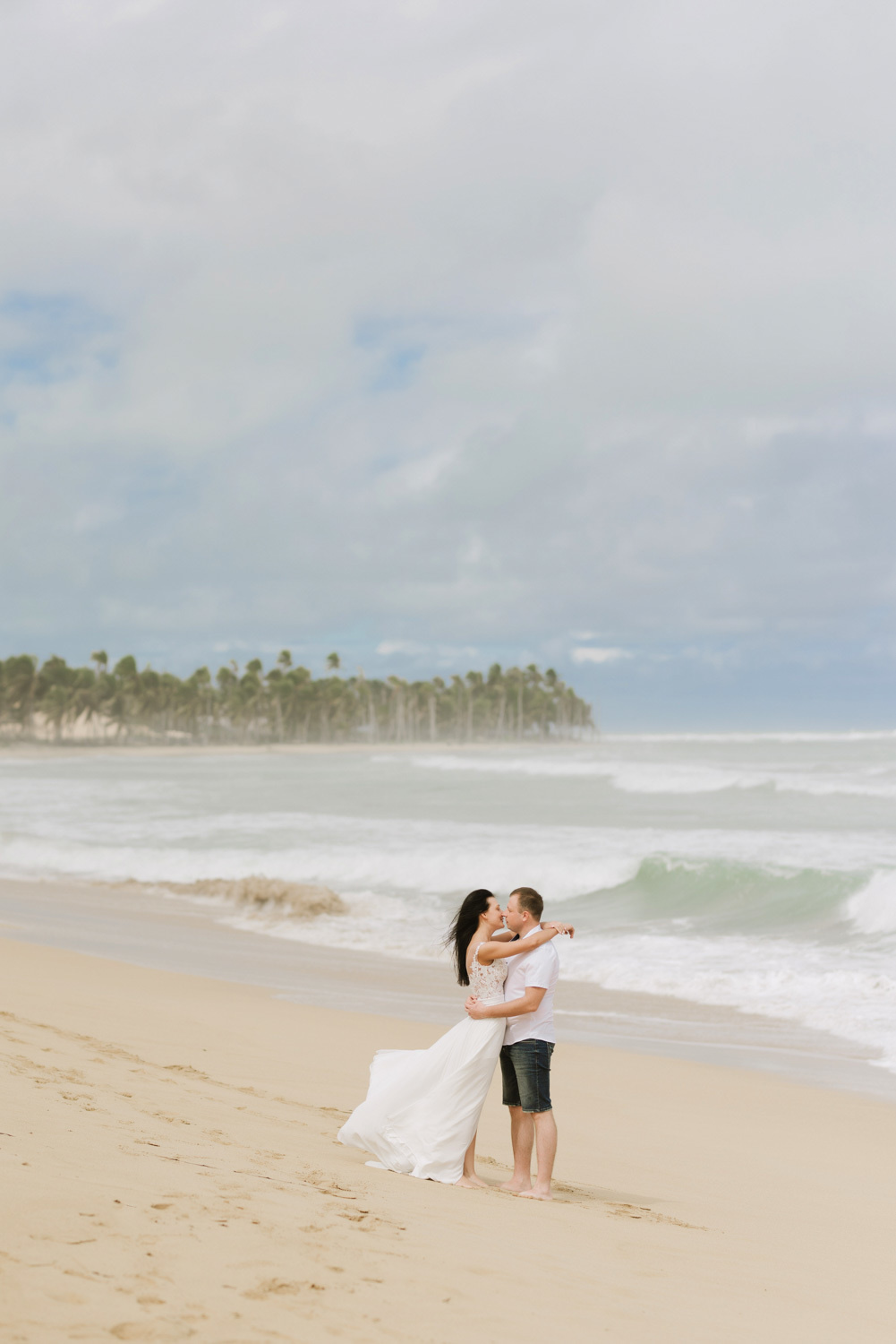 The groom hugs and kisses the bride in a flowing white dress on the background of the sea bay.