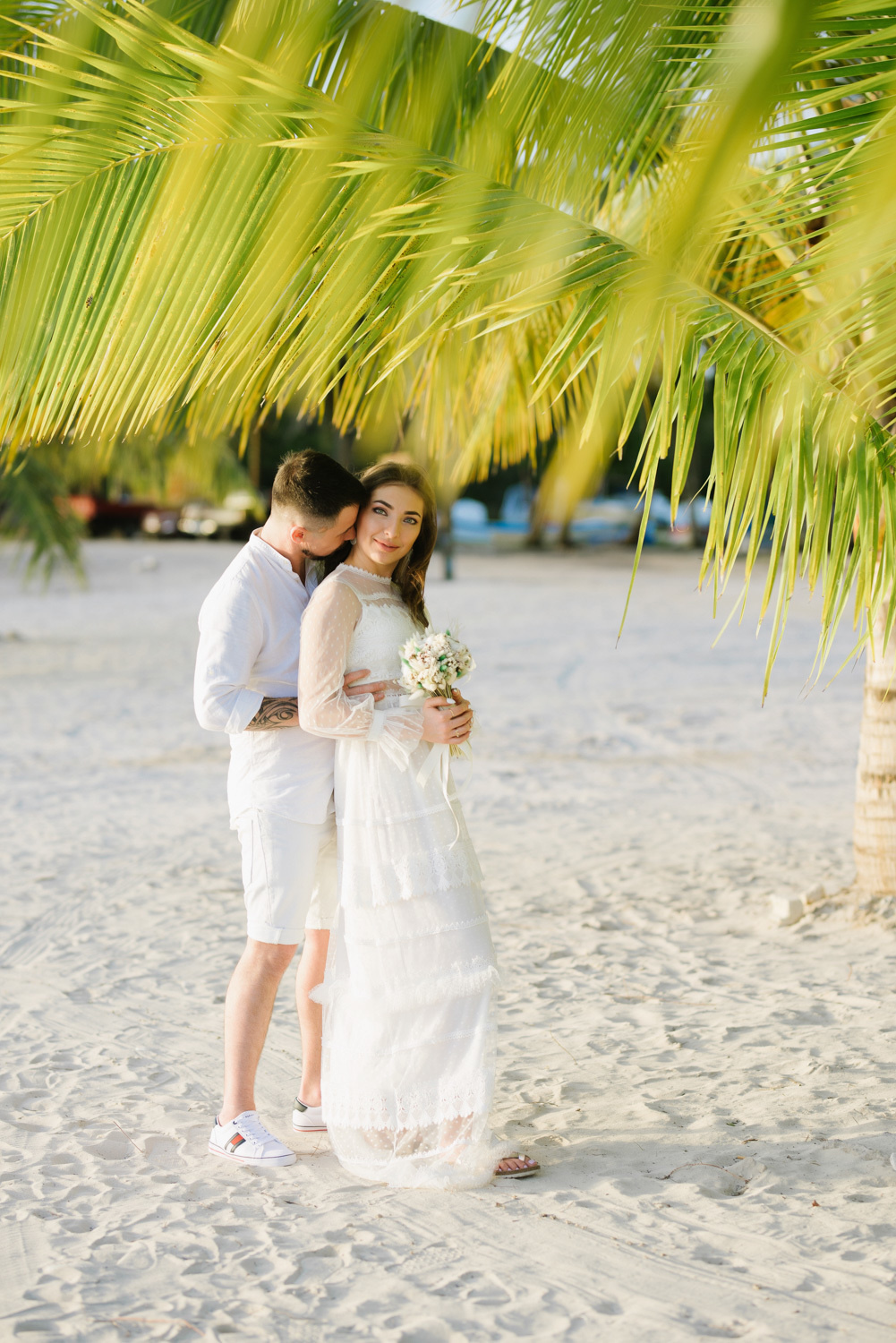 Under a palm tree on the white sand, the groom embraces the bride.
