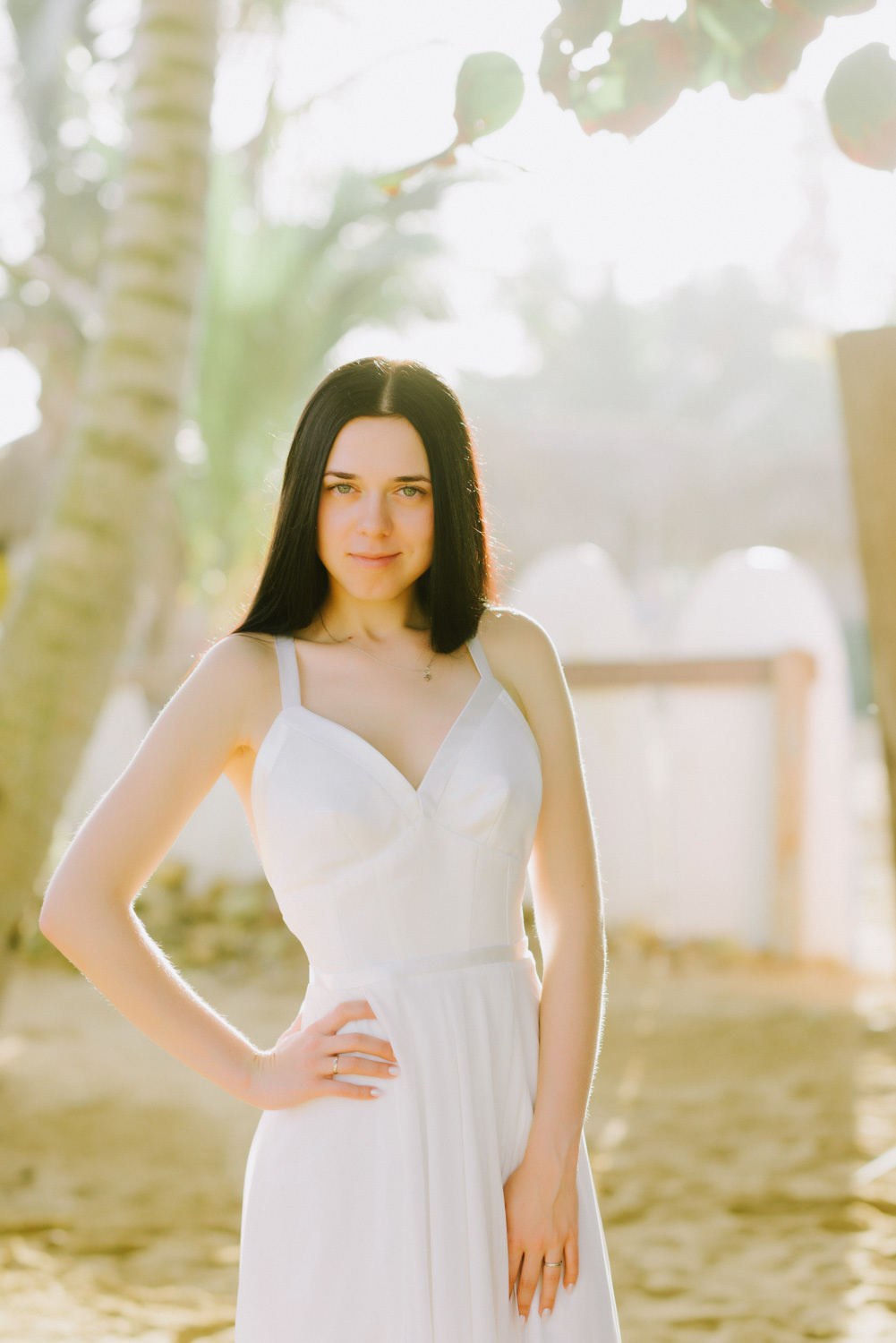 The bride's face with straight long dark hair on the background of a beach with palm trees, illuminated by bright sunlight.