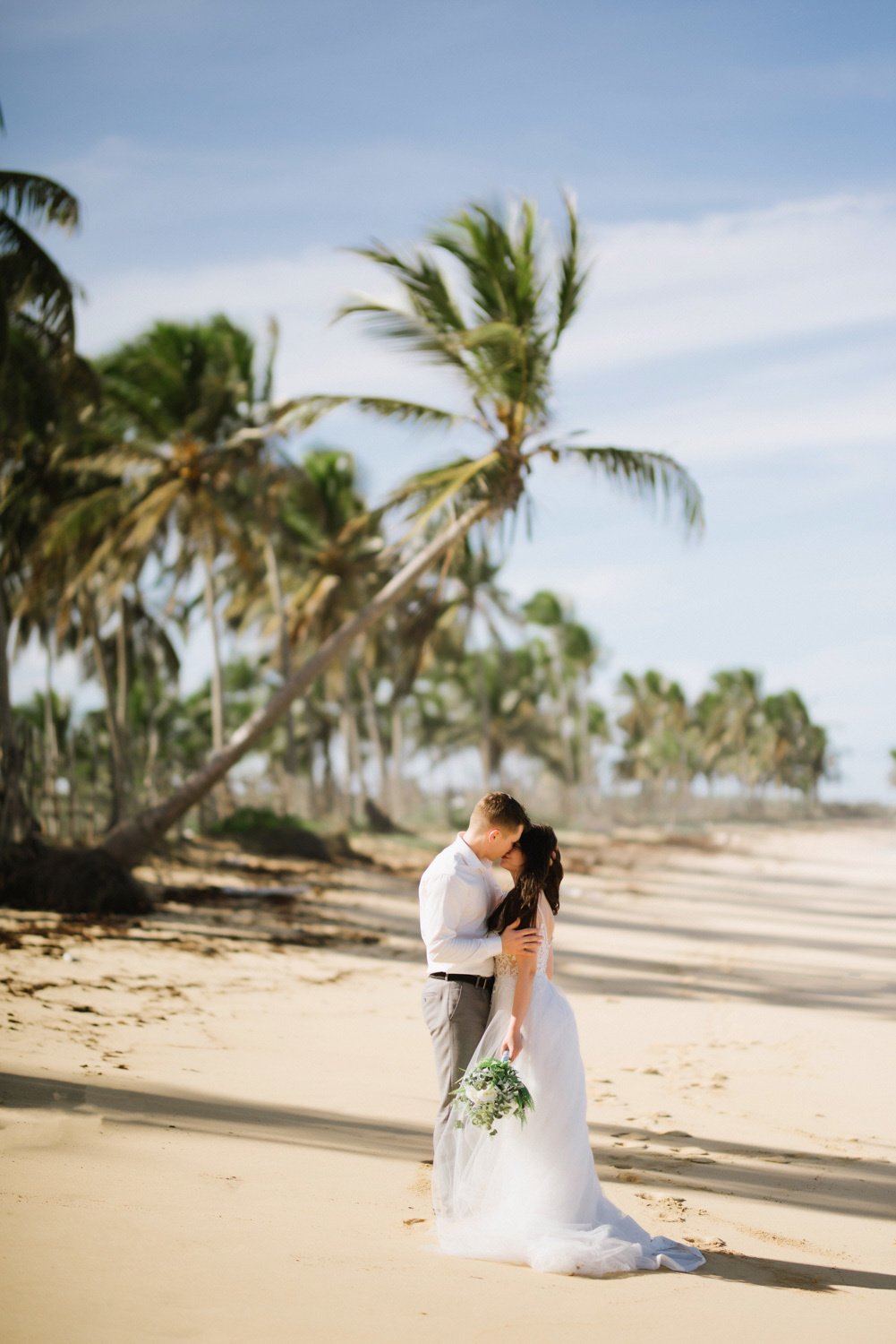 On the beach with palm trees, the groom kisses the bride.