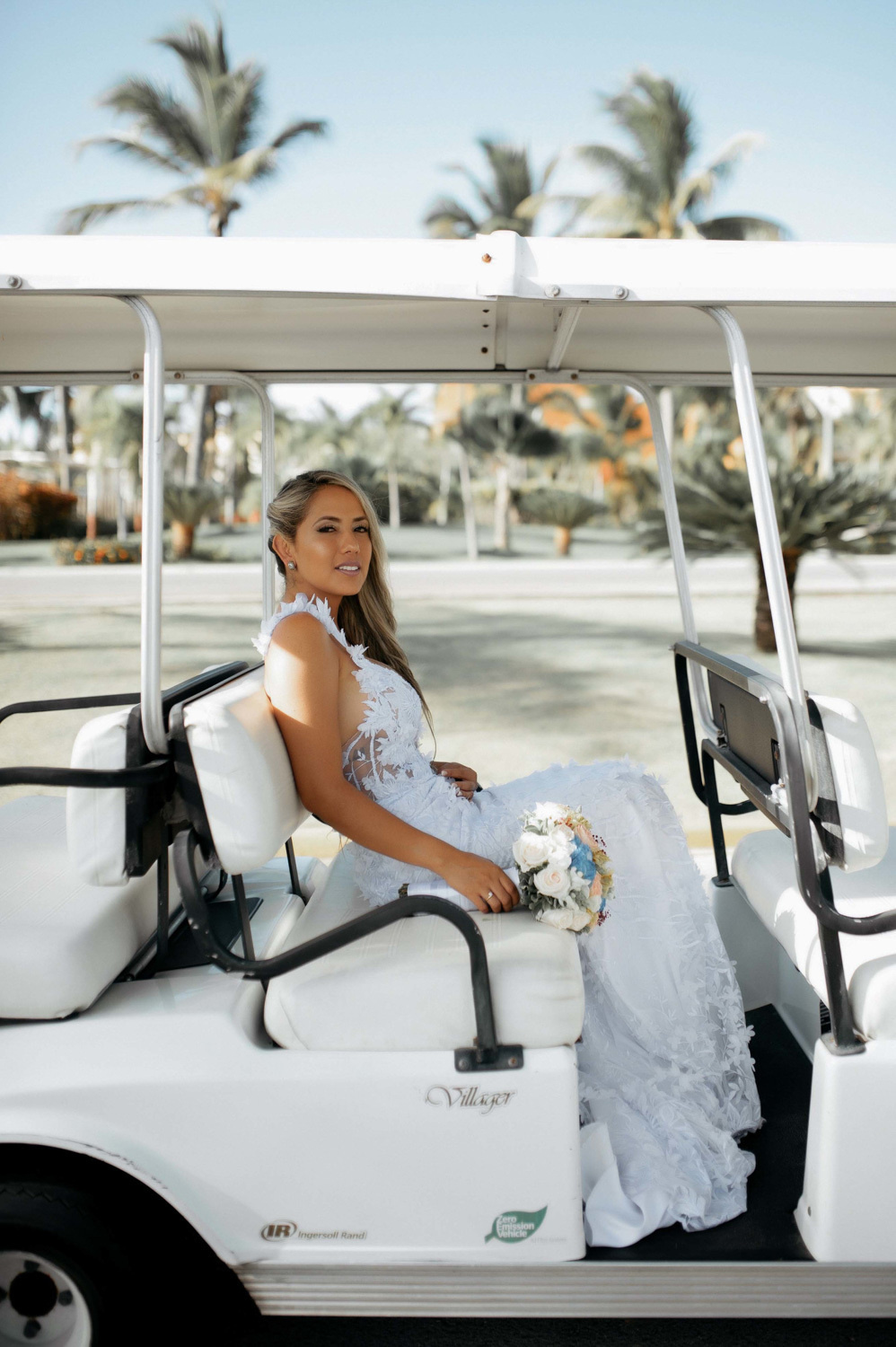 A bride in a wedding dress sits in a summer open car. On the seat next to her is a bouquet of flowers.