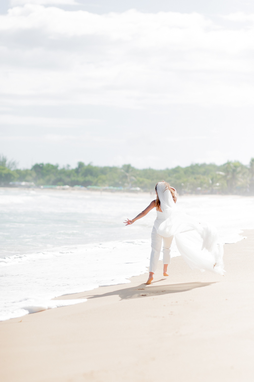 The bride has her back turned and is looking out to sea. White trousers are visible from under the fluttering dress.