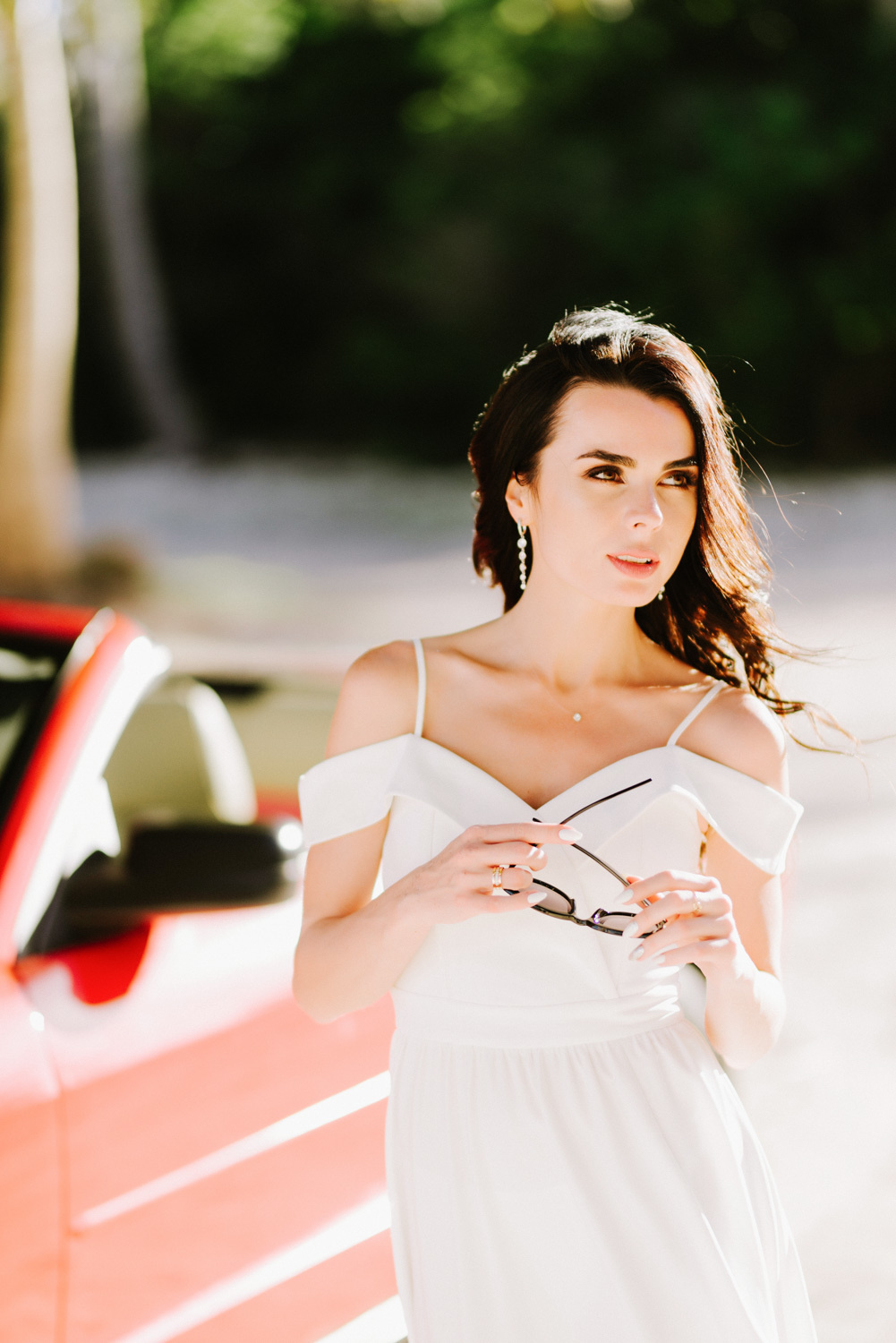 Невеста у красной машины держит в руках очки от солнца на фоне зеленого массива.