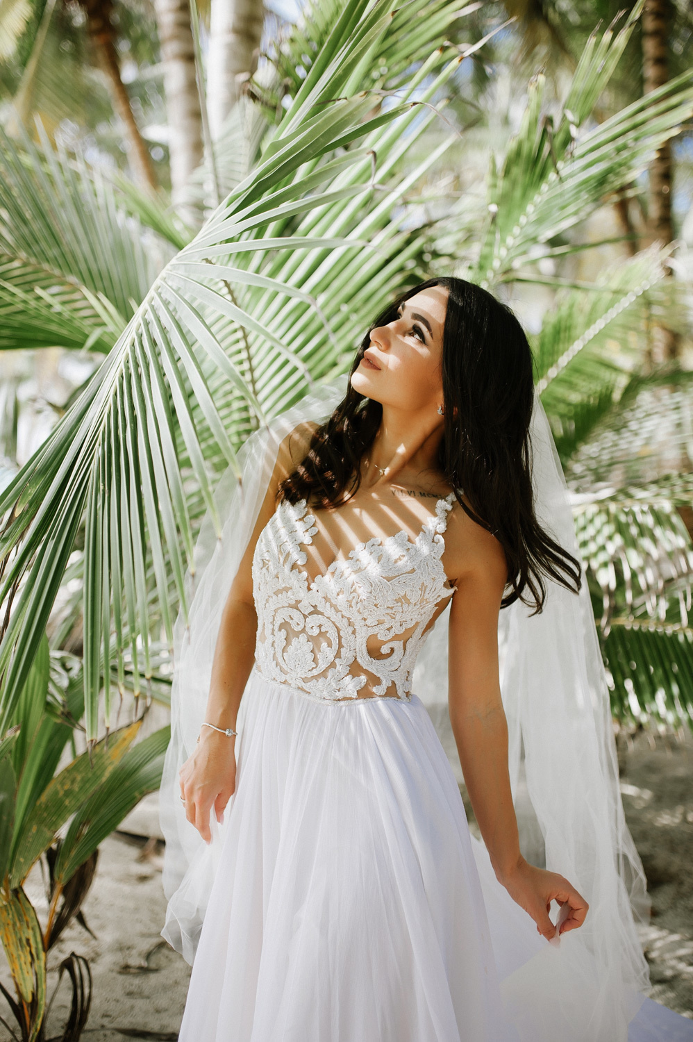 The bride looks at the sun, the rays of which illuminate her through a huge palm leaf.