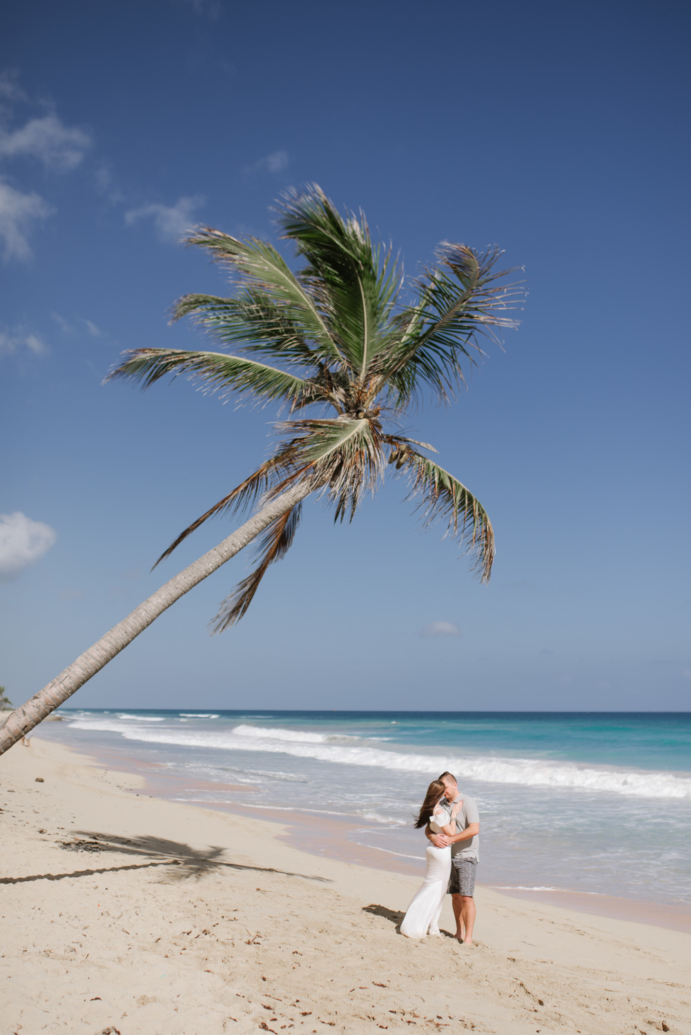 On a sandy beach with a tilted palm tree, the bride and groom stand with their arms around each other against the background of the sea.