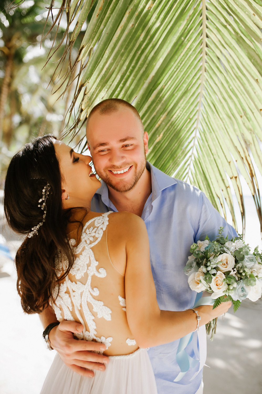 A dark-haired bride in a wedding dress with a bouquet of flowers in her hand is hugged by the groom with a short haircut and beard on the background of a palm tree.