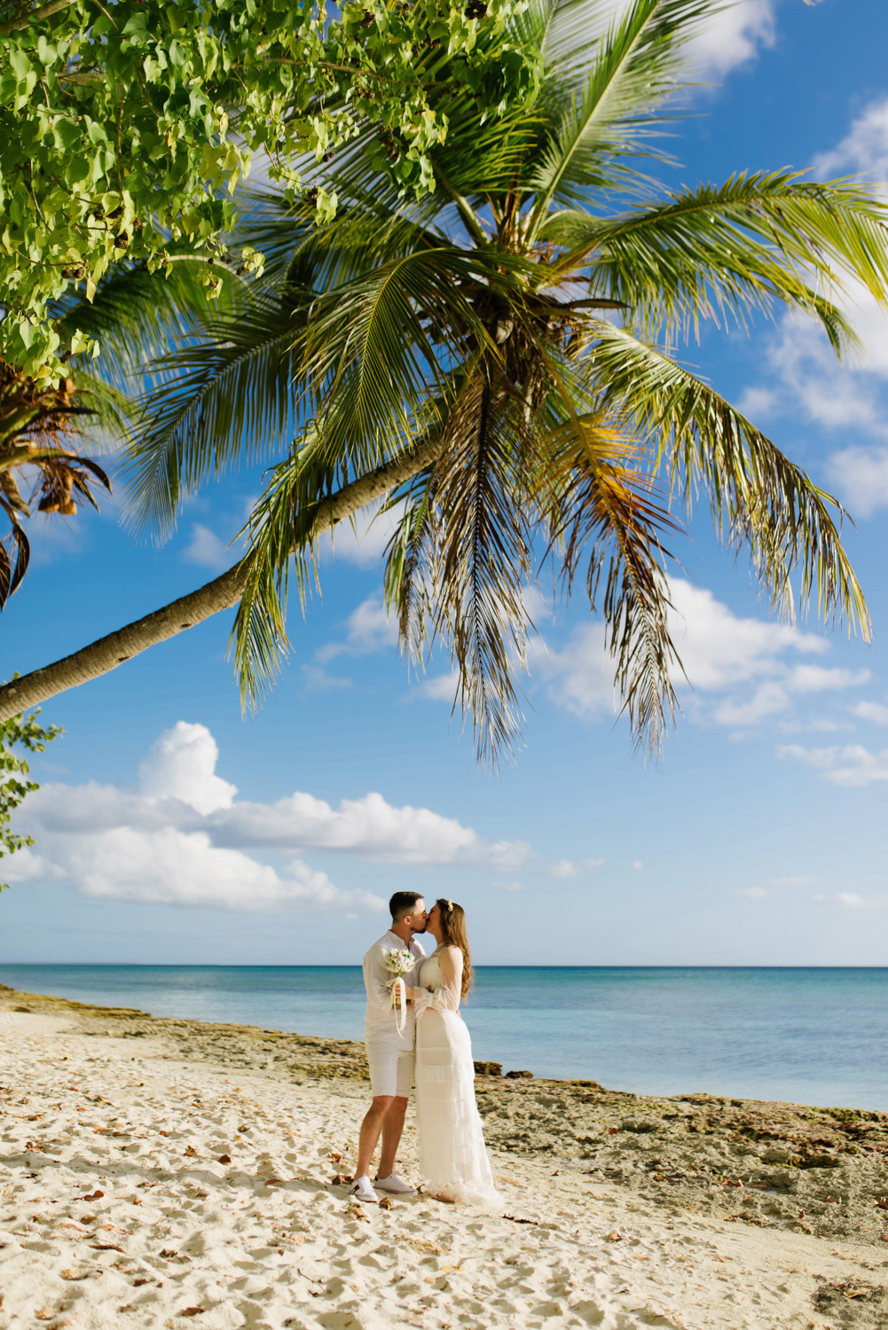 Under a tall palm tree on the beach near the sea, the bride and groom kiss against the background of a blue sky with white clouds.