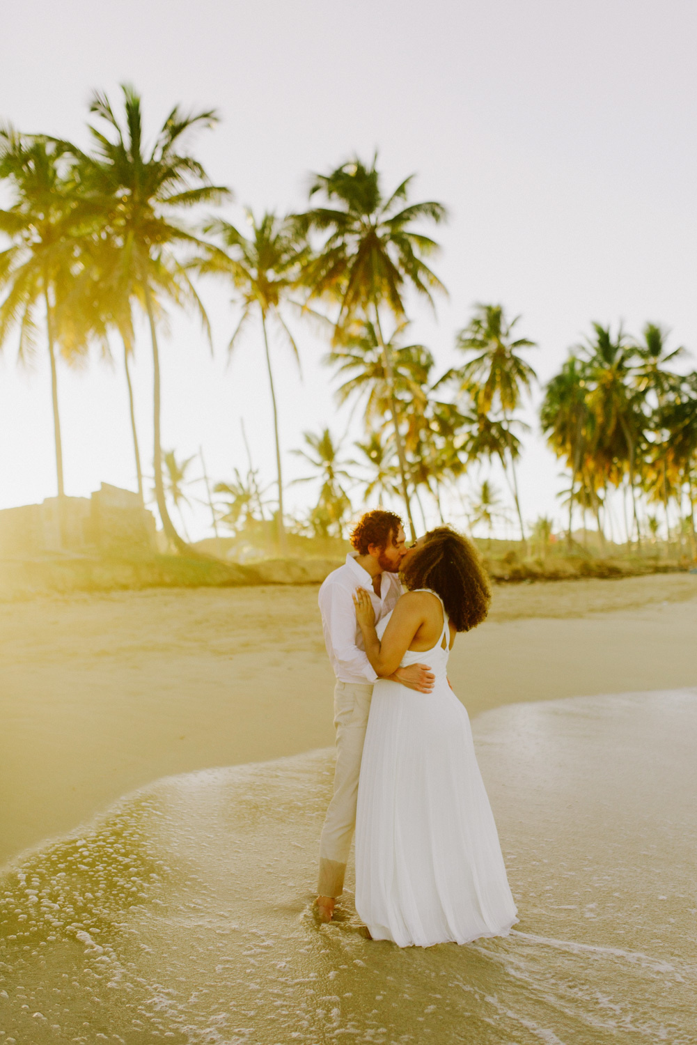 Стоя босиком в морской воде, жених и невеста целуют друг друга на фоне песчаного берега с пальмами.