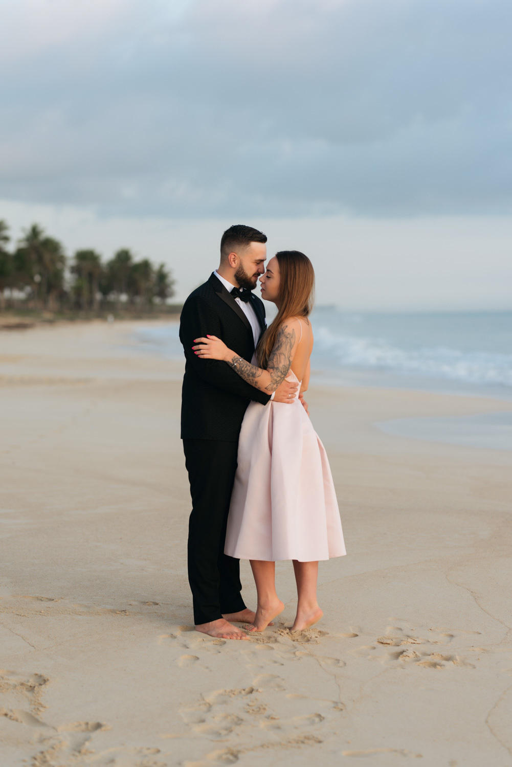 A dark-haired man with a short haircut, beard and mustache in a dark suit, white shirt with a bow tie embraces a girl with loose brown hair in a pink short puffy dress on the sandy beach.