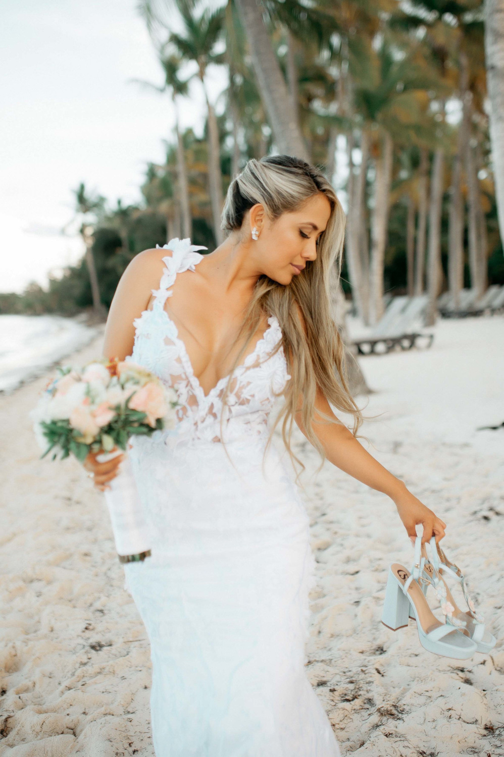 The bride walks along the sandy beach with huge palm trees in the background. She has a bouquet of flowers in one hand and a pair of sandals in the other.