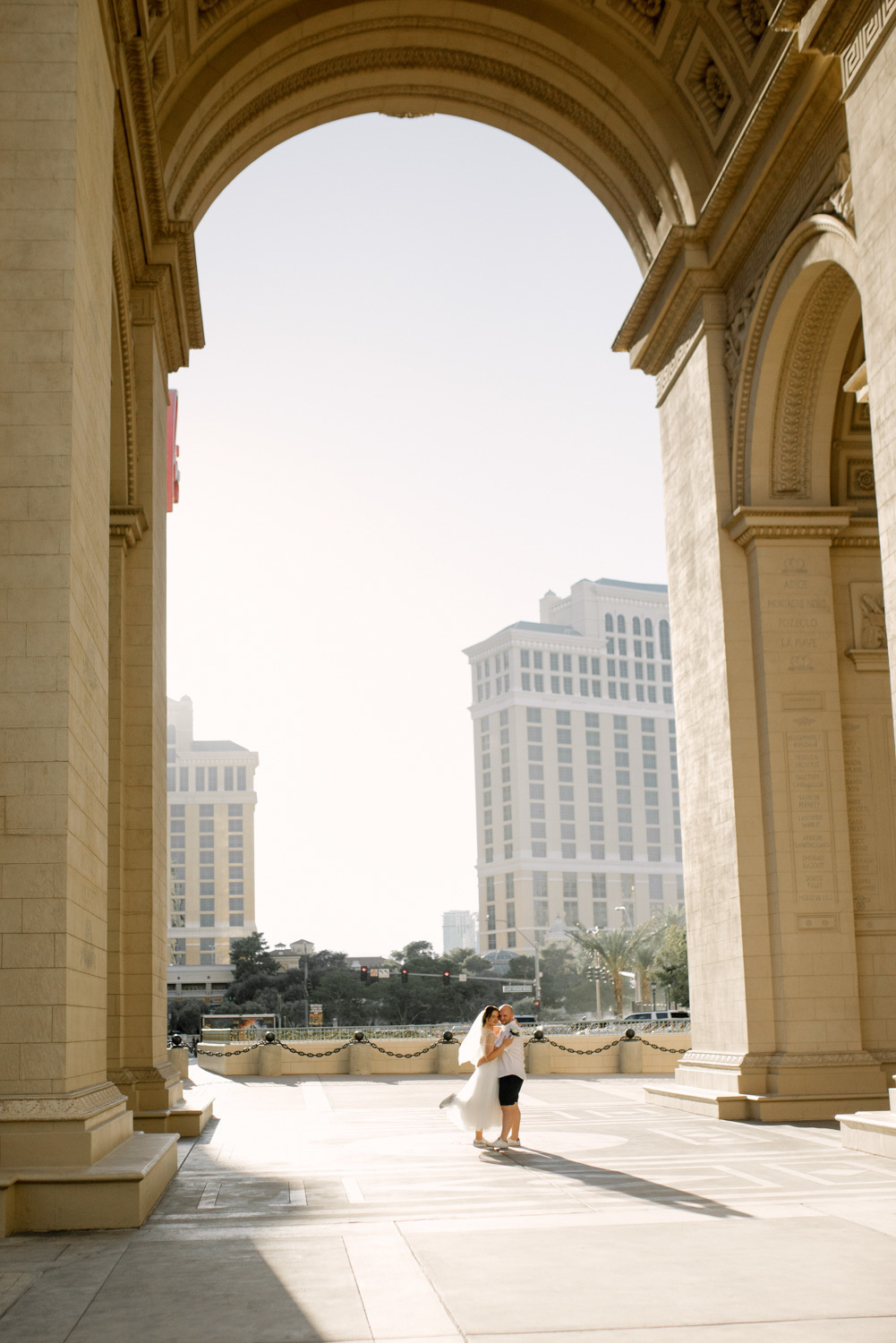 В центре огромной арки на фоне белоснежных зданий и зеленого сквера жених обнимает невесту.