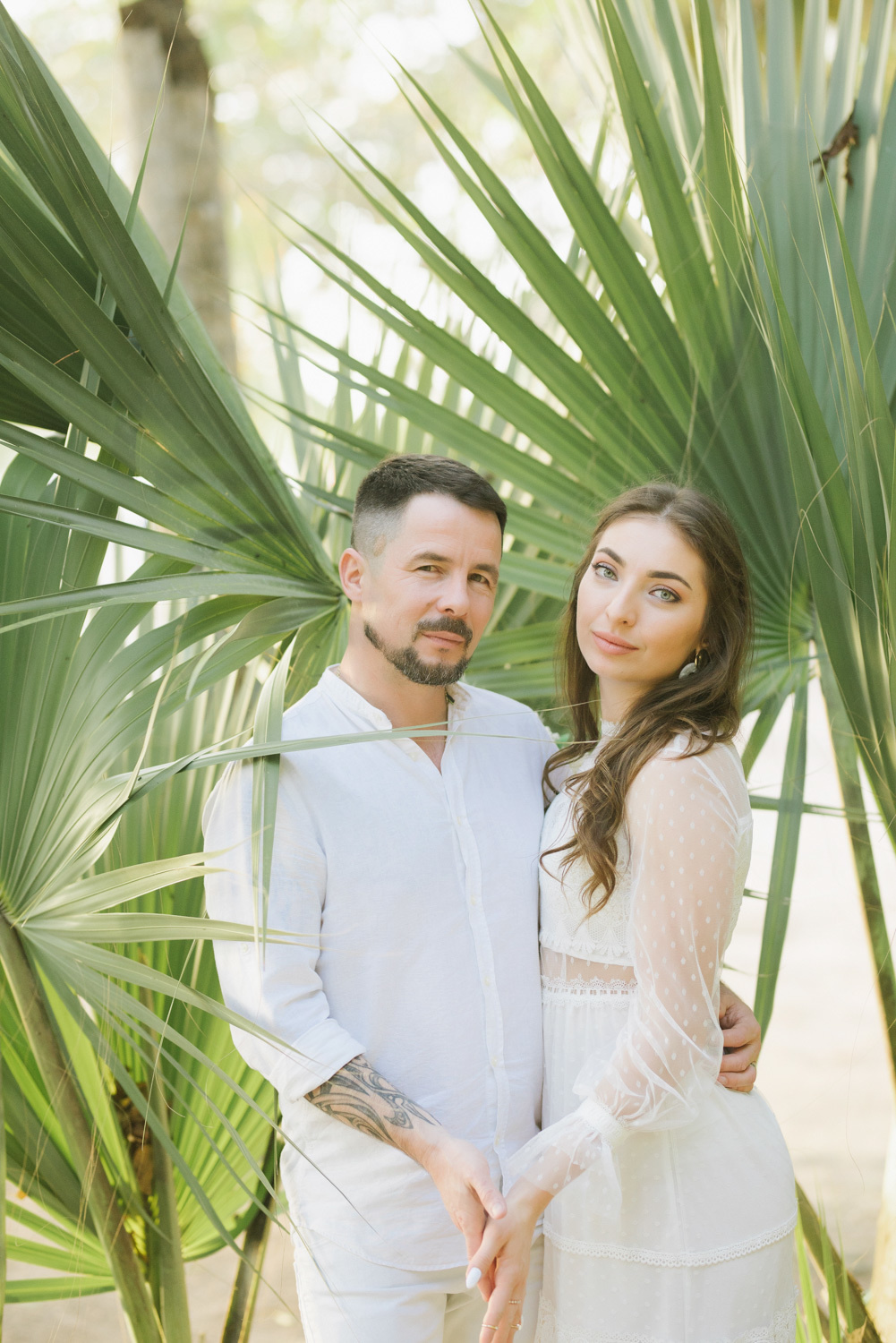 The bride and groom hold hands and look at the photographer against the background of huge palm leaves.