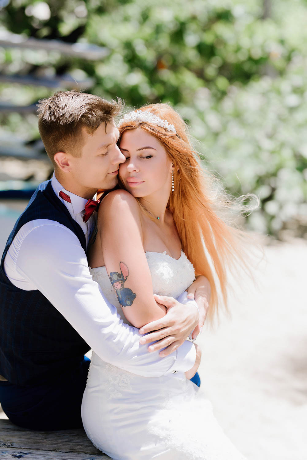 The groom embraces the bride from behind against the background of a blurry green landscape.