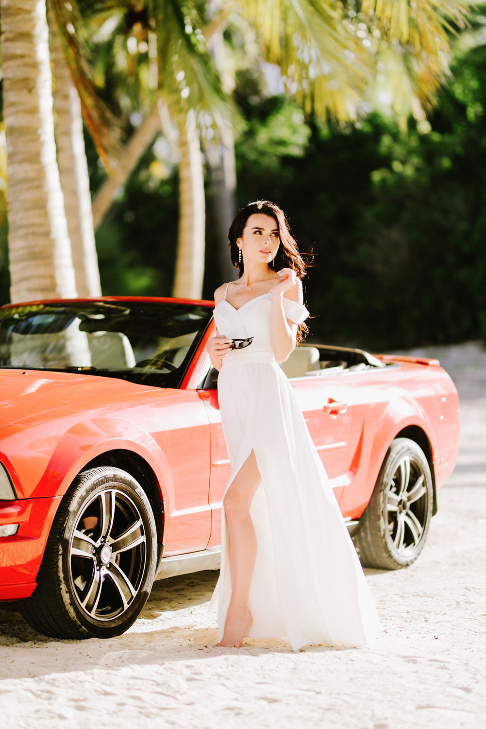 Невеста в длинном белом платье у красного автомобиля на фоне пальм.
