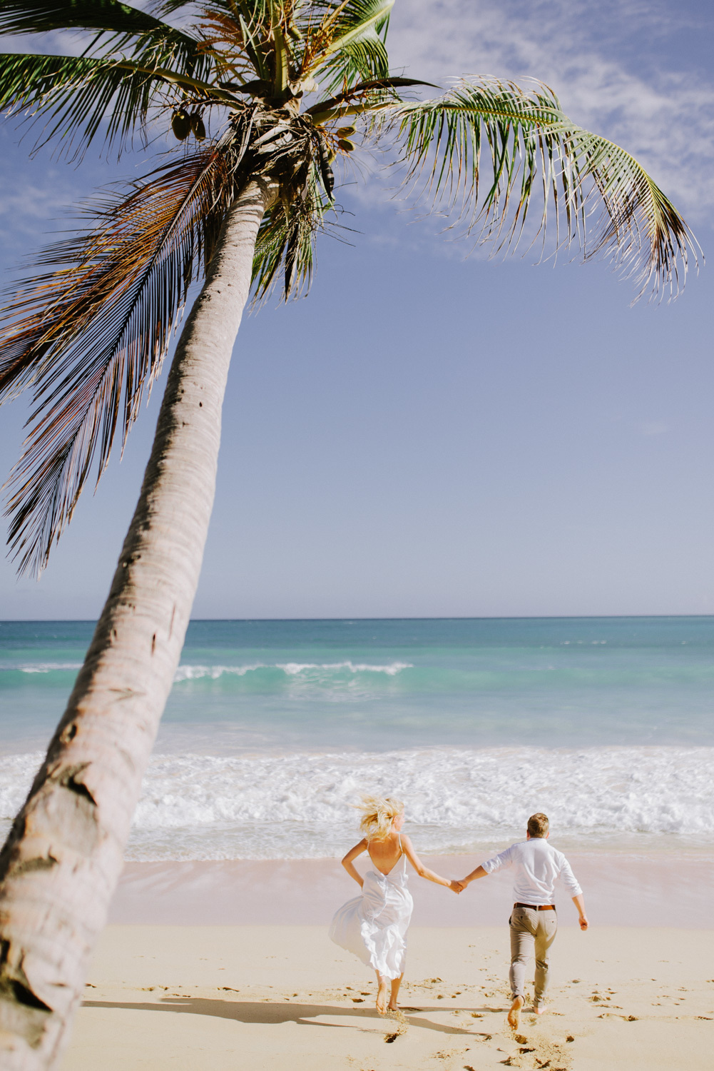 Past a tilted palm tree, the bride and groom walk hand in hand to the sea, leaving footprints in the sand.