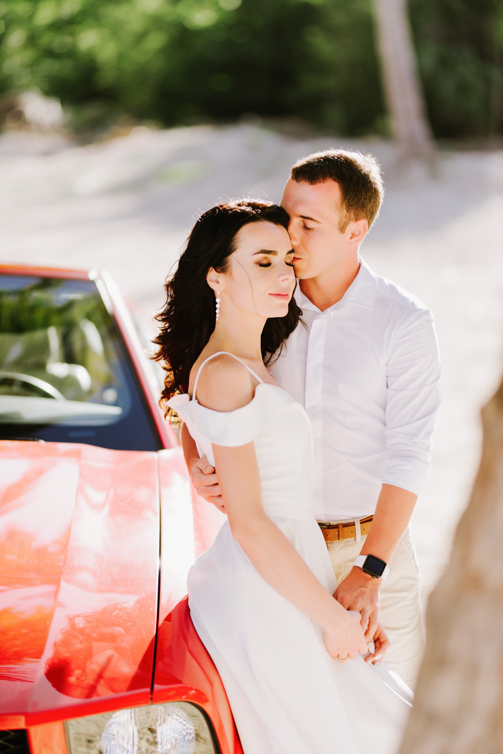 Невеста с женихом обнимаются у красного автомобиля на фоне пальм.