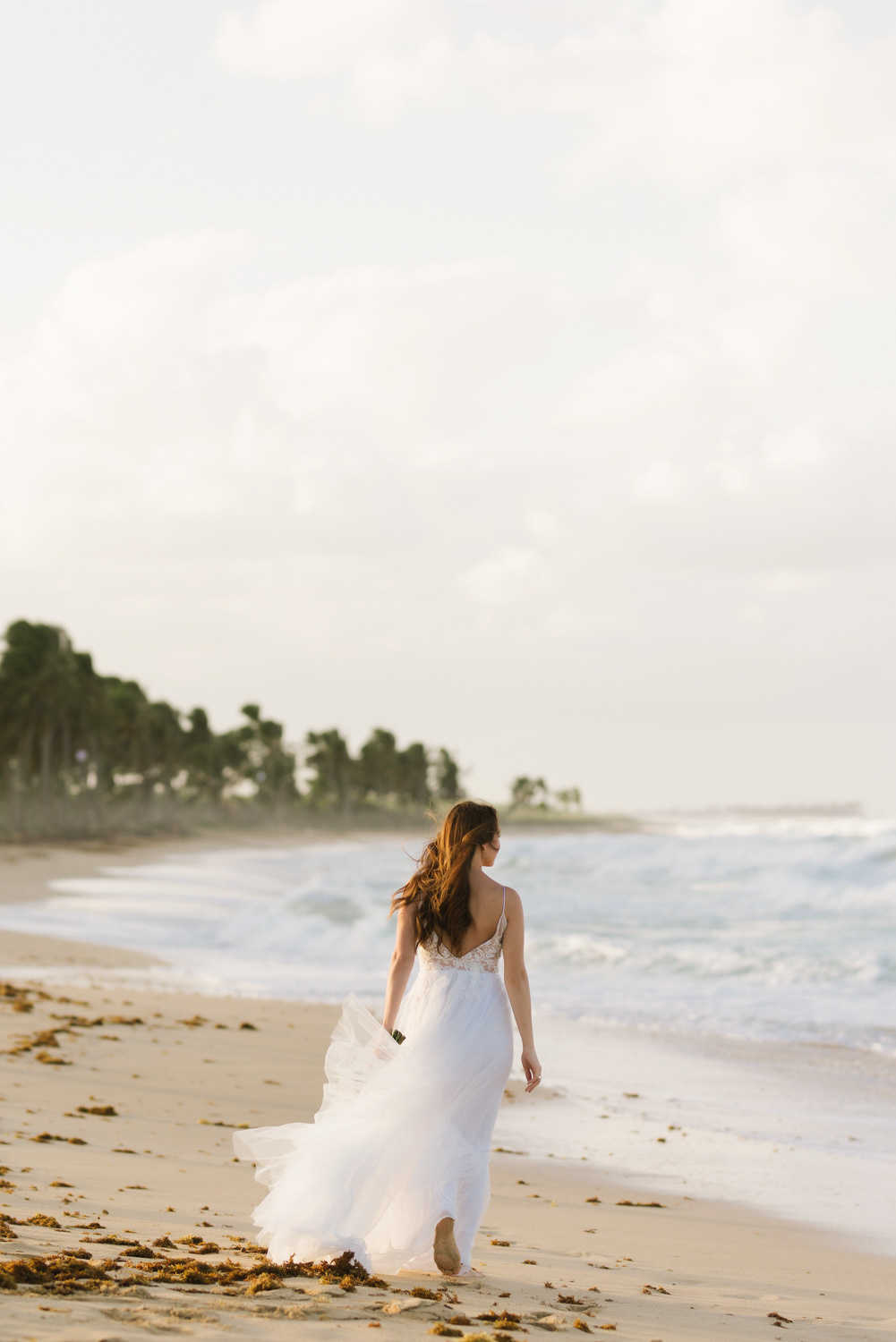 a bride in a wedding dress walks on a sandy beach barefoot, rear view